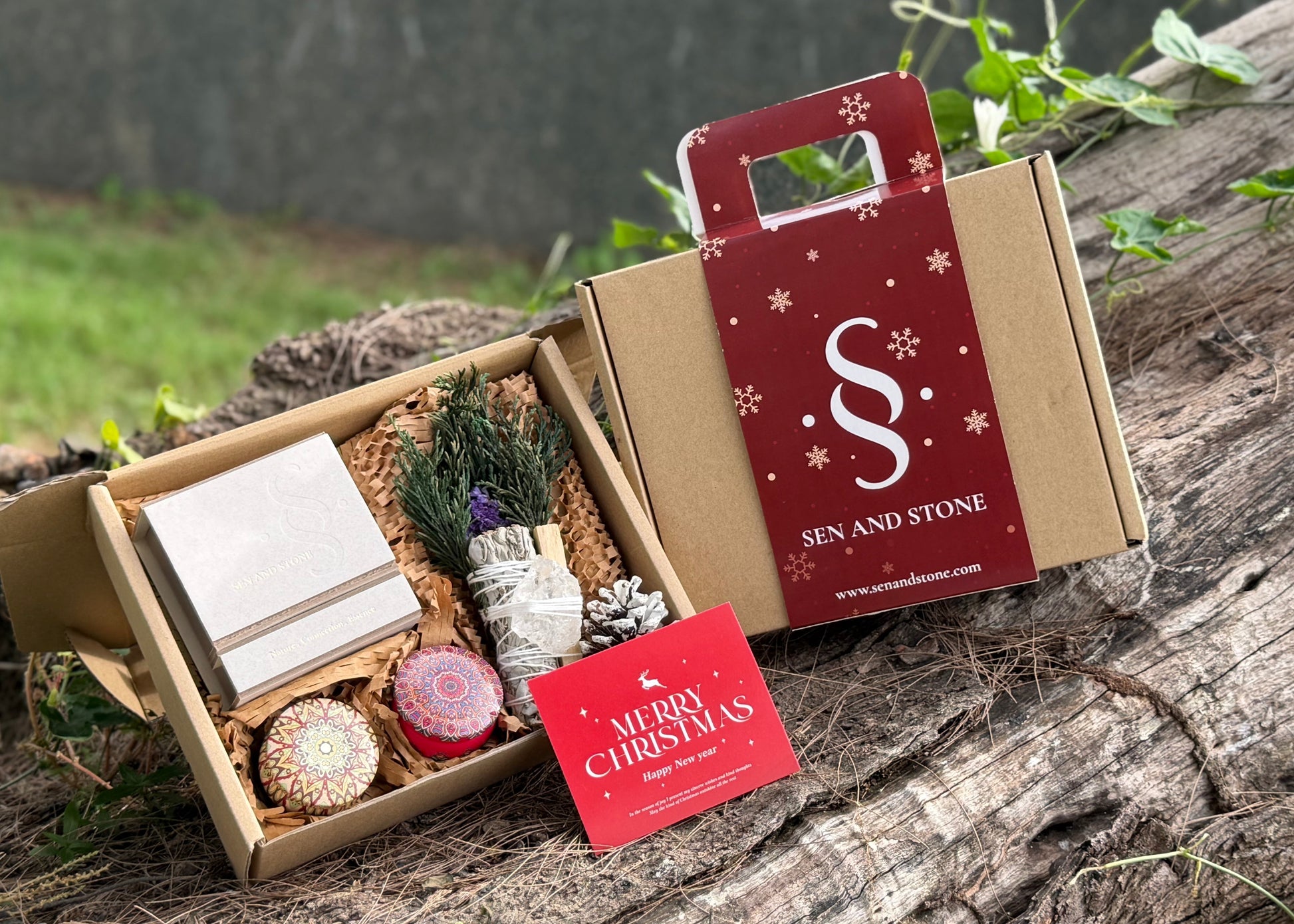 Gift box with decorative items and a branded card on a wooden surface outdoors.